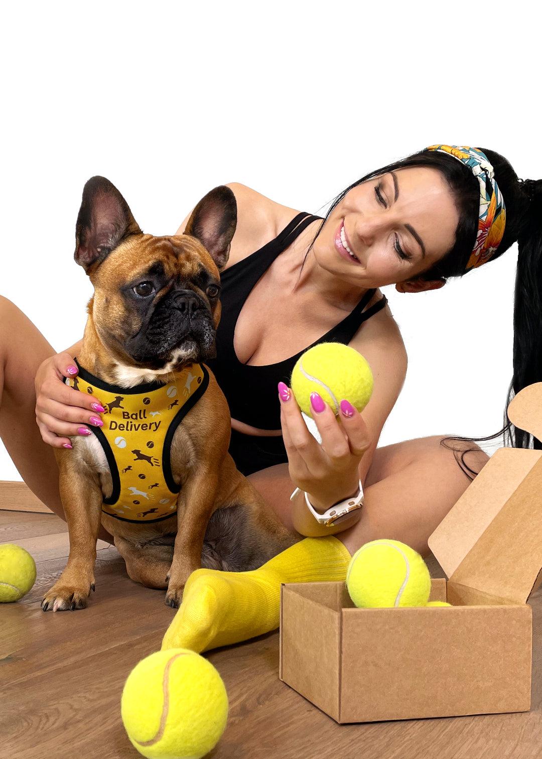 French bulldog wearing a ball delivery design harness, with depicted illustrations of running dogs and tennis balls, in dark brown and white on a yellow background. The dog sits in front of her human mom, who's leaning a little sideways to see dogs reaction and smiles, while the dog is looking at a ball she's holding. There's also an open cardbox with more tennis balls in and outside.