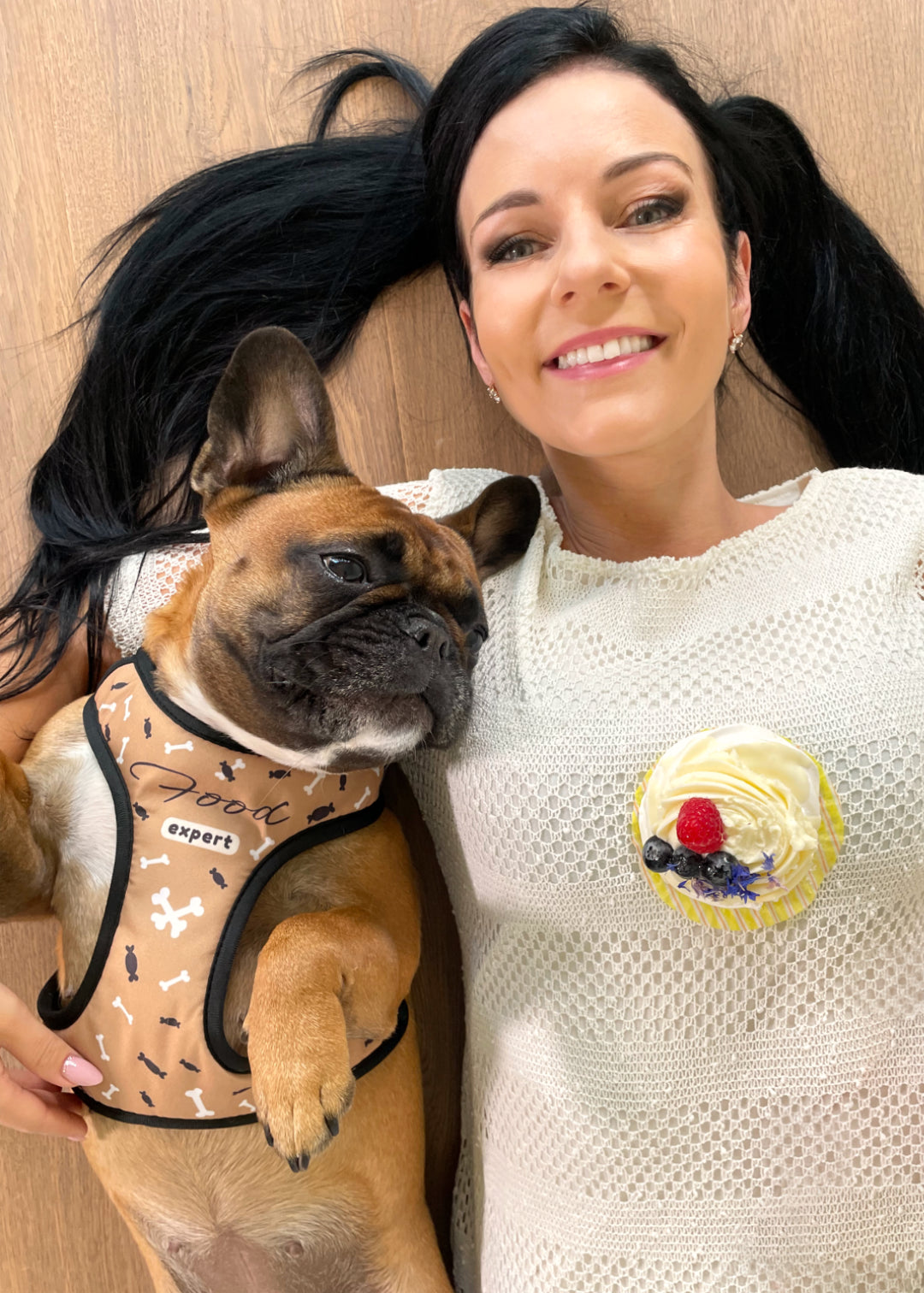 French bulldog with her human mom laying on wooden floor, the picture is taken from upside of them, both are enjoying the chill lay on the floor, showcasing the food expert design harness, while Vicky is looking at a cupcake that's on top of her moms chest.