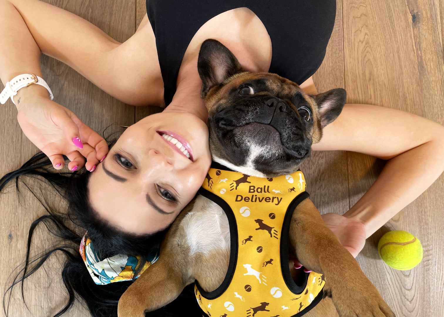 French bulldog wearing yellow themed ball delivery design harness, laying on her back head to head with a young woman bot smiling looking upwards.