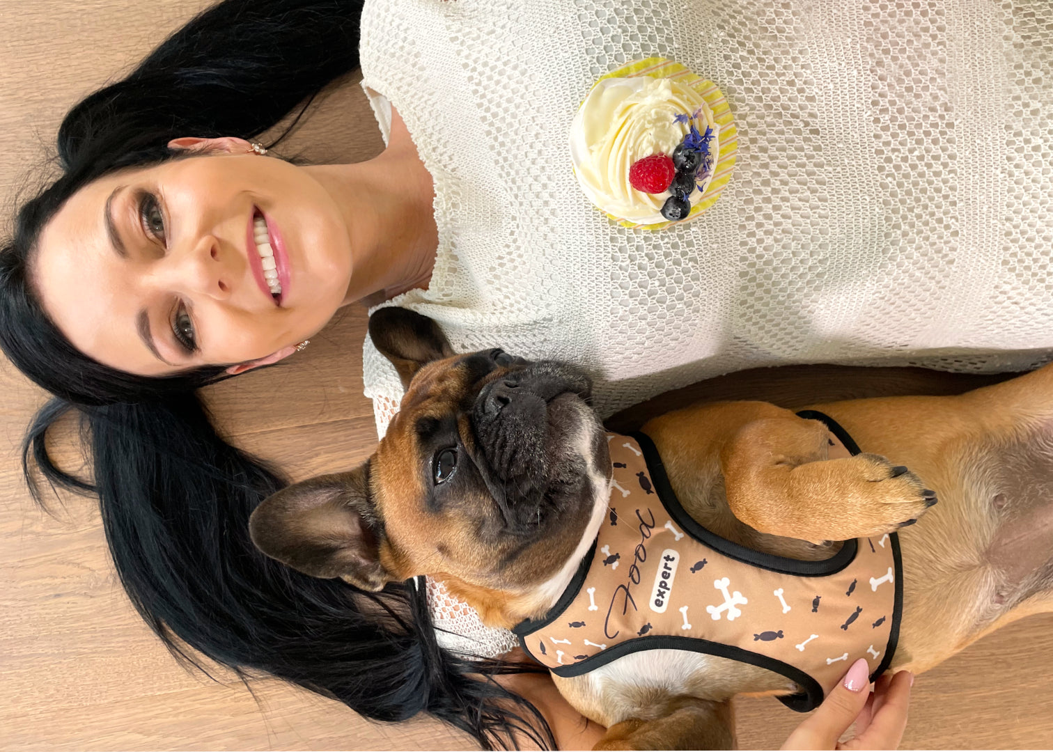 French bulldog and her human mom laying on wooden floor both happy while the dog is wearing sand colour themed food expert design harness and is looking at a cupcake that's on her moms chest.